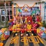 Group of children in bright traditional costumes posing for a janmashtami festival photo at a school stage with colorful umbrellas and marigold decorations in the scene